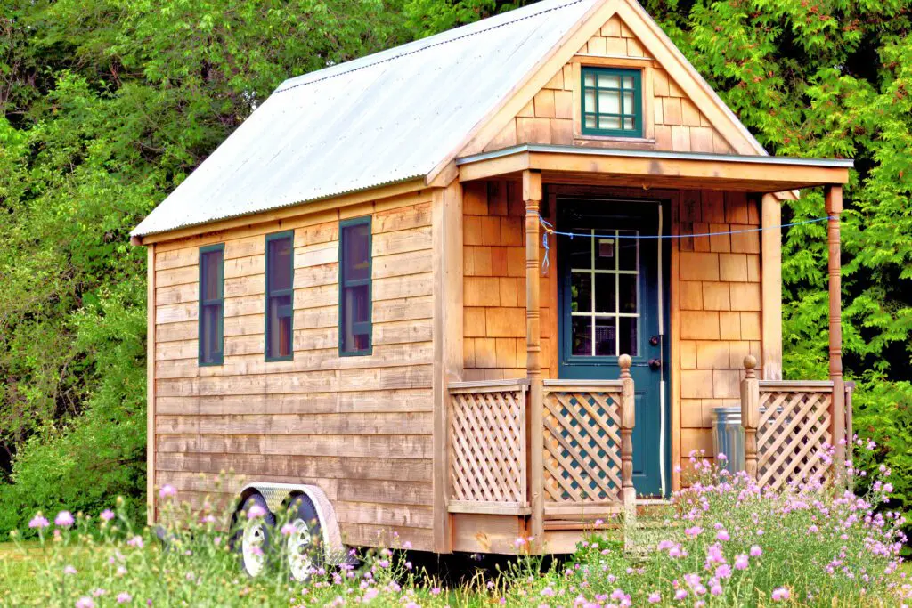 Exterior view of a tiny house on wheels with wooden siding, surrounded by greenery and wildflowers.