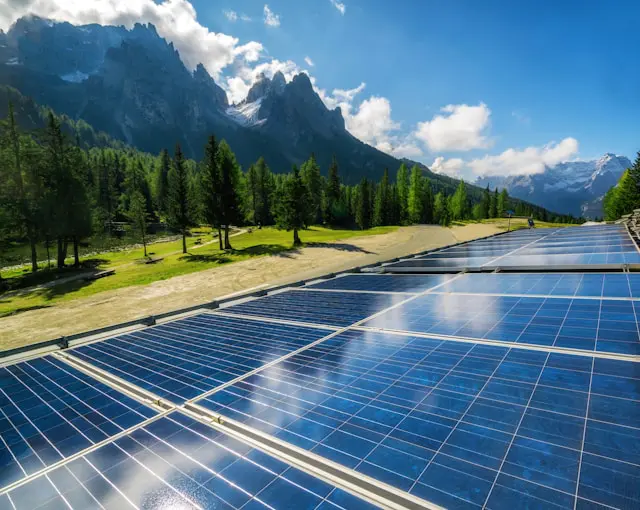 Solar panels in a scenic mountain landscape with green trees and a clear blue sky.