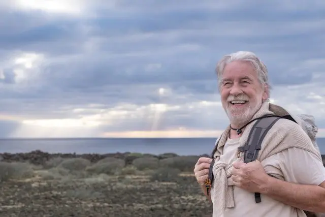 An elderly man with a grey beard, smiling broadly, wearing a backpack, standing against a backdrop of a cloudy sky and calm ocean.
