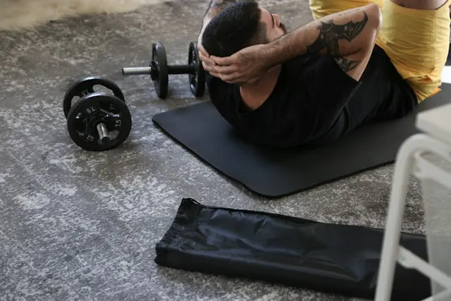 Man performing abdominal crunches next to dumbbell and mat in a home gym setting.