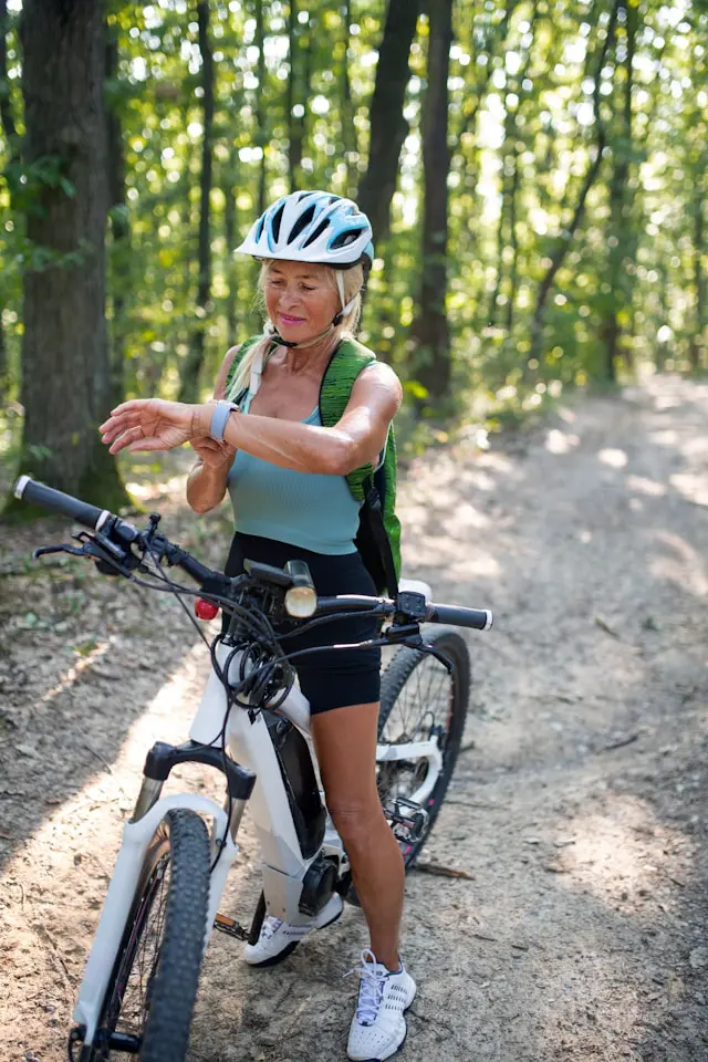 Woman in athletic wear checking her smartwatch while standing with her ebike on a forest trail.