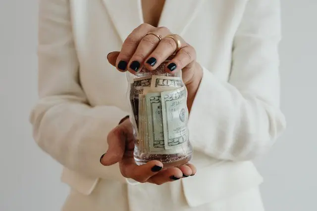 A woman in a white blazer holding a glass jar filled with US dollar bills, showcasing financial savings or donations.