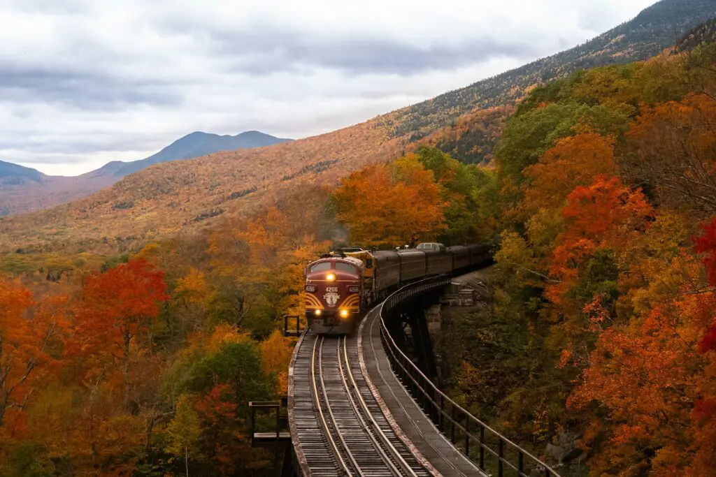 A vintage-style train crossing a curved mountain bridge with bright orange and yellow fall foliage, set against misty mountains in the background.