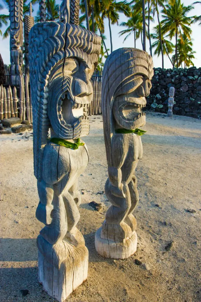 Two intricately carved wooden statues with traditional Hawaiian designs, set against a backdrop of palm trees and a stone wall.