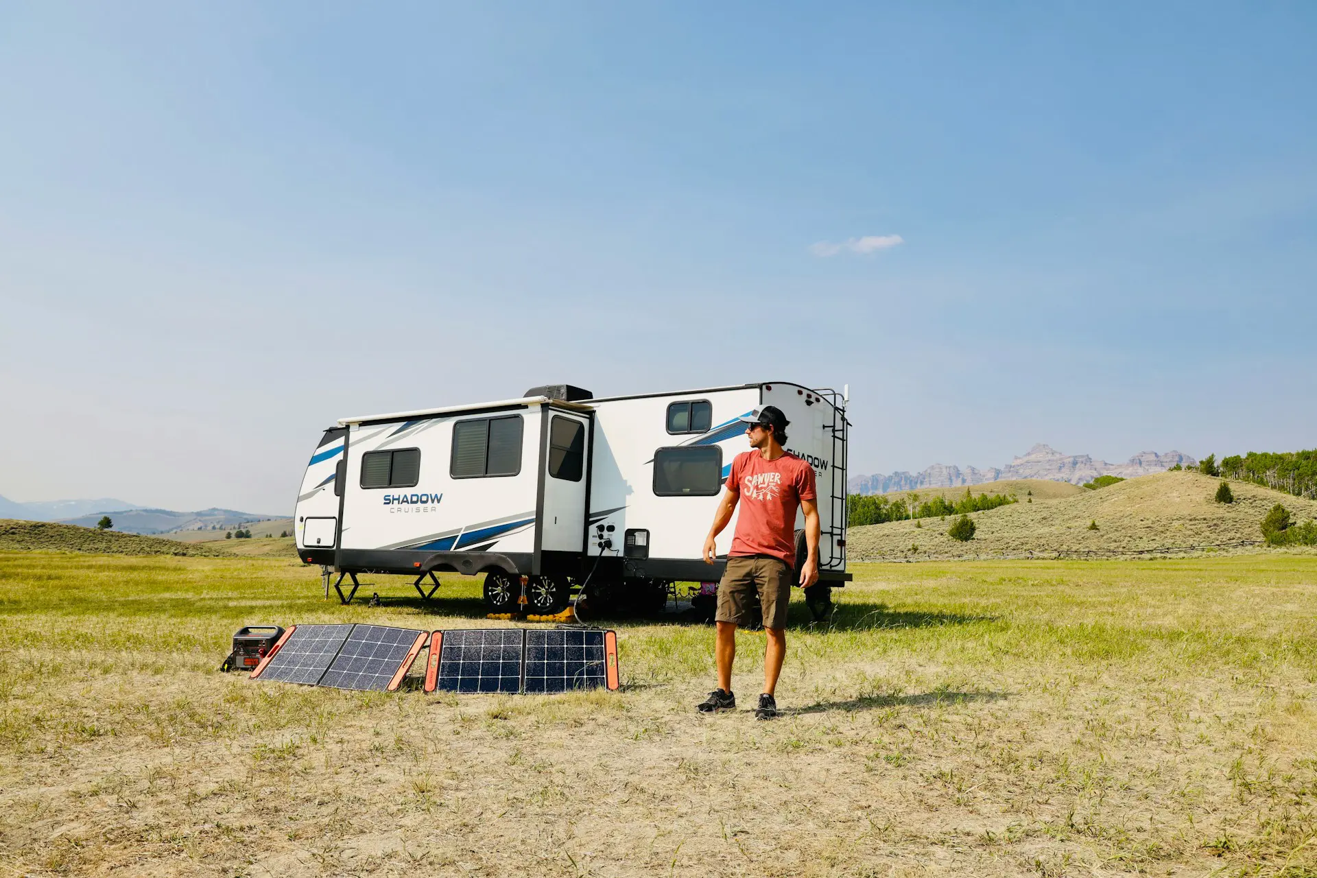 Man in red shirt setting up solar panels next to a white and blue RV in a sunny field with distant mountains visible in the background.