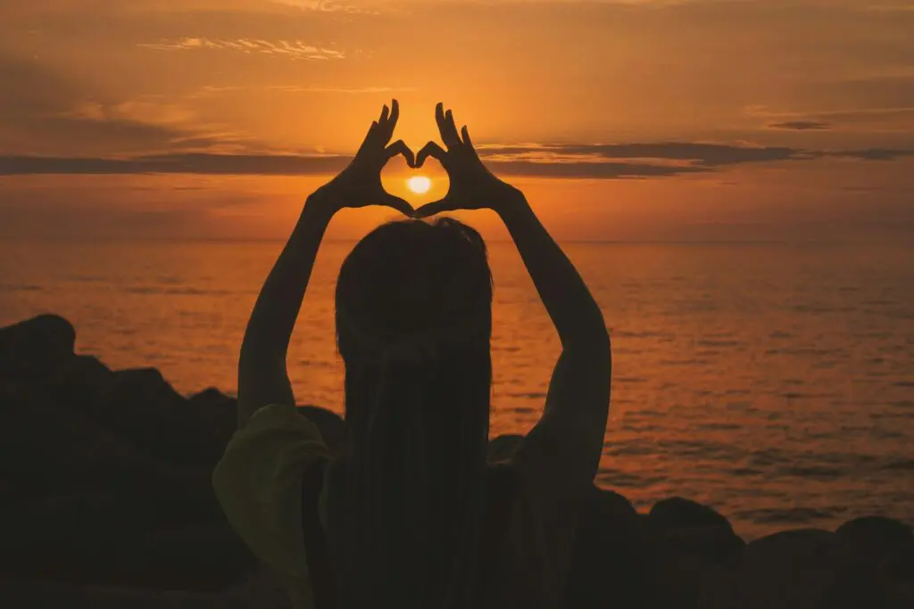 Silhouette of a woman forming a heart shape with her hands against a sunset over the ocean.