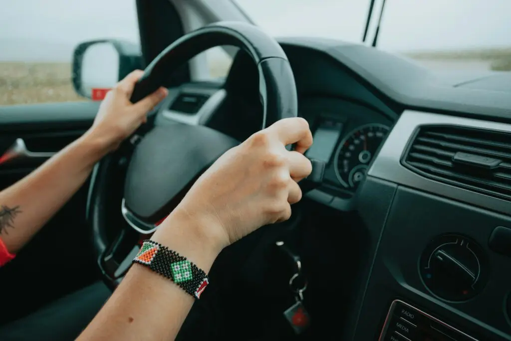 Close-up of hands on a car's steering wheel, wearing a beaded bracelet, driving during the day.