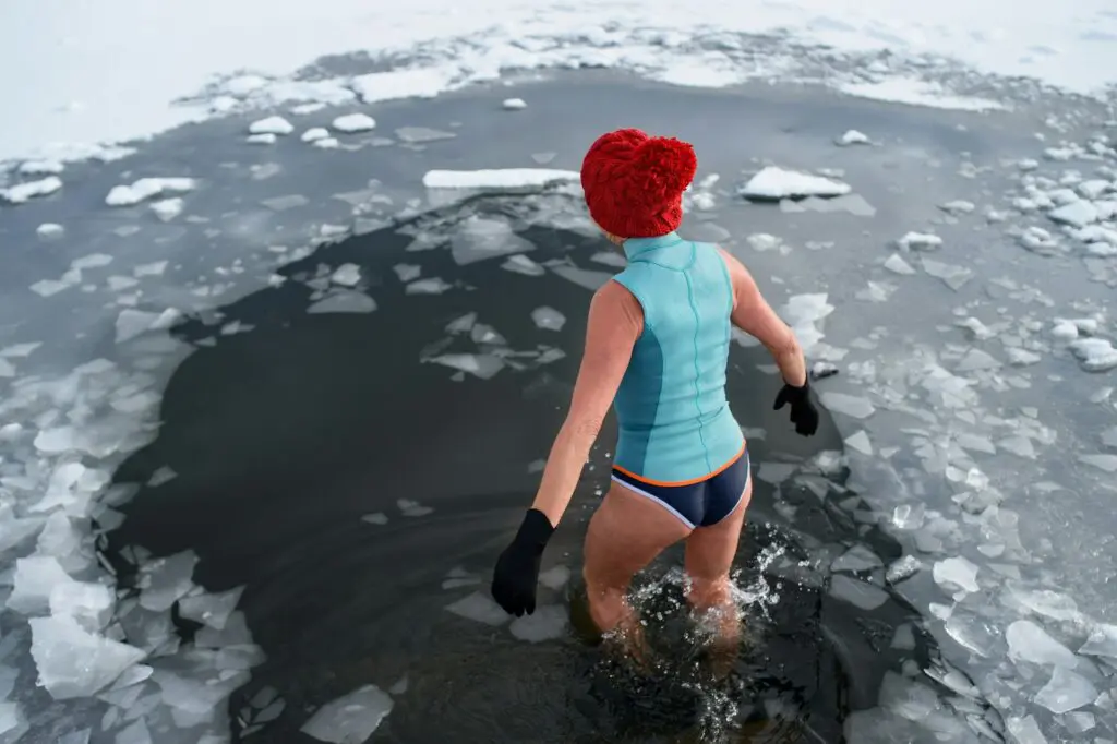 A woman in a red knit cap confidently enters an icy water hole in winter, demonstrating resilience and cold exposure therapy.