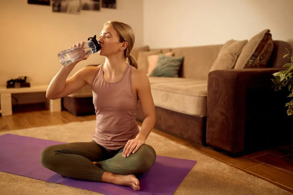 Woman in yoga pose on a purple mat, drinking water from a clear bottle, in a serene living room.