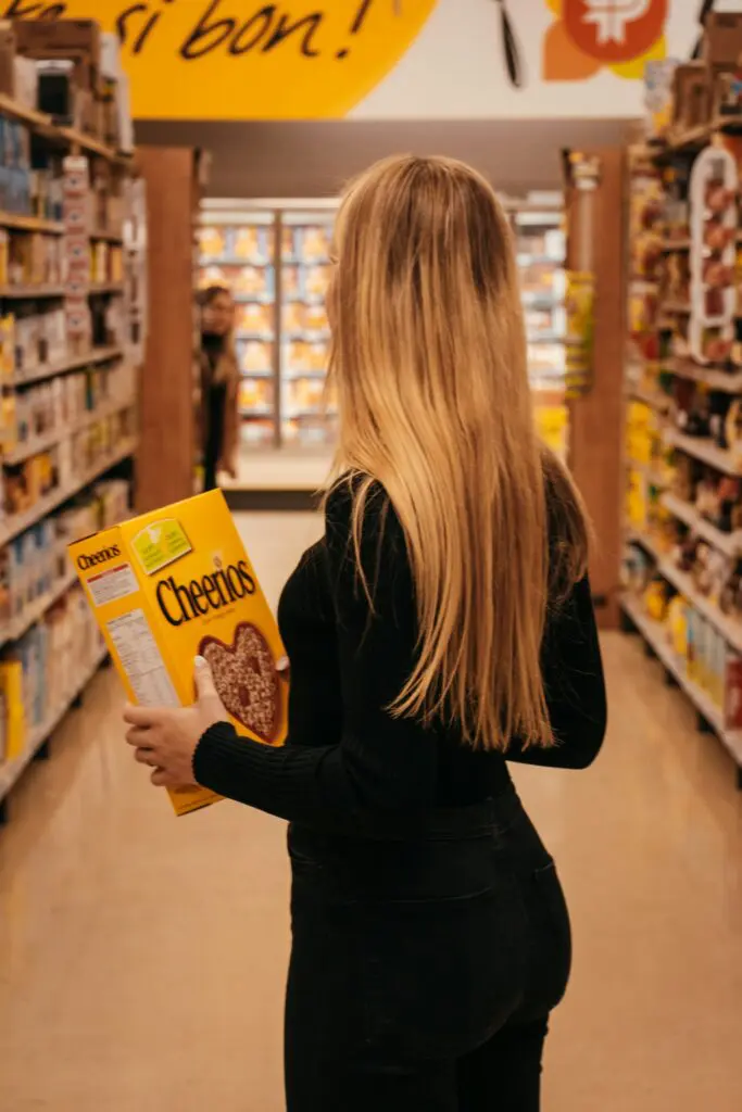 A woman with long blonde hair holding a large box of Cheerios while browsing in a grocery store aisle.