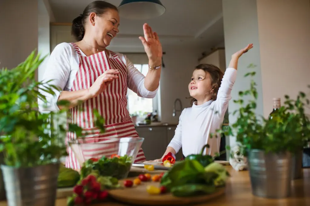 A joyful scene in a home kitchen where a grandmother in a striped apron and her young granddaughter happily prepare a fresh salad together.