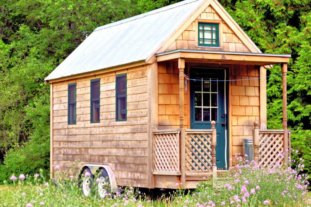 Exterior view of a tiny house on wheels with wooden siding, surrounded by greenery and wildflowers.