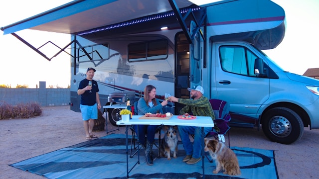 A group of RV newbies enjoying a meal outside their RV, featuring essential RV gear such as folding chairs, a table, an awning for shade, and two dogs.