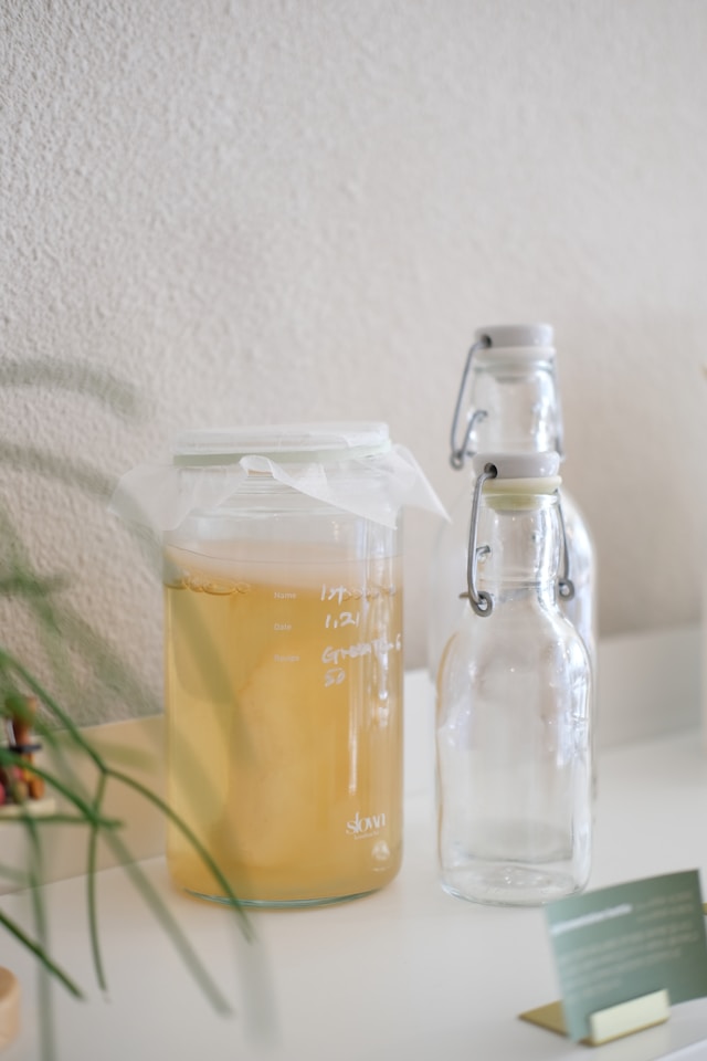 A glass jar containing a SCOBY in the process of fermenting kombucha, accompanied by two empty glass bottles with swing-top caps on a white surface.