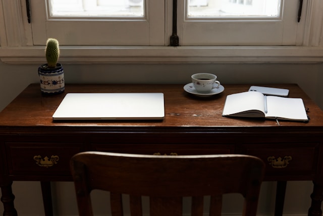 Minimalist wooden desk with a closed laptop, open notebook, cup of coffee, and a small cactus plant near a window