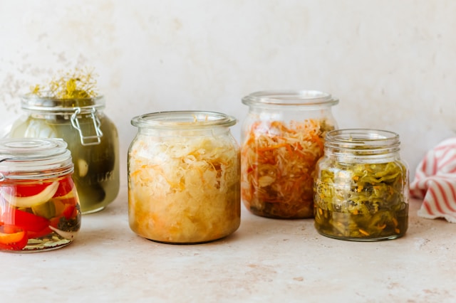 Various jars of homemade fermented vegetables, including pickles, sauerkraut, and kimchi, displayed on a light-colored surface.