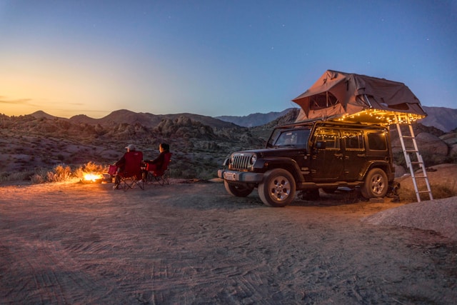 Jeep with a rooftop tent illuminated by string lights at sunset, with two people sitting by a campfire in a desert landscape.