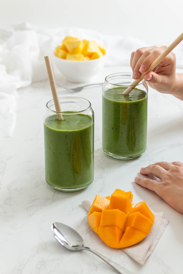 Two glasses of green smoothie with bamboo straws on a marble countertop, with a sliced mango and bowl of diced pineapple, highlighting how to heal your gut naturally.