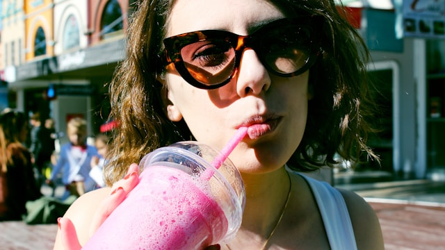 A woman wearing sunglasses and drinking a strawberry smoothie through a straw in an outdoor setting.