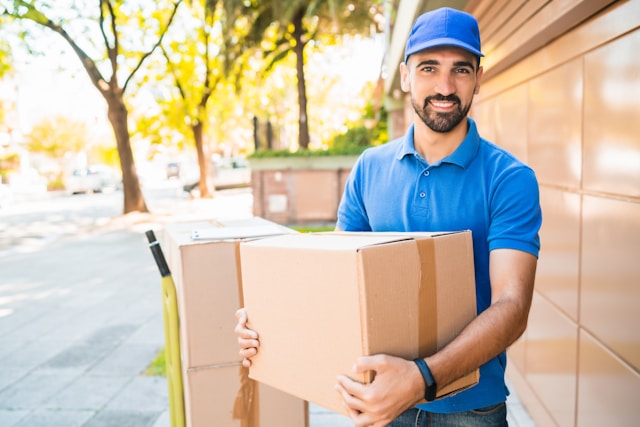 A mail delivery person in a blue uniform holding a cardboard package outdoors on a sunny day.