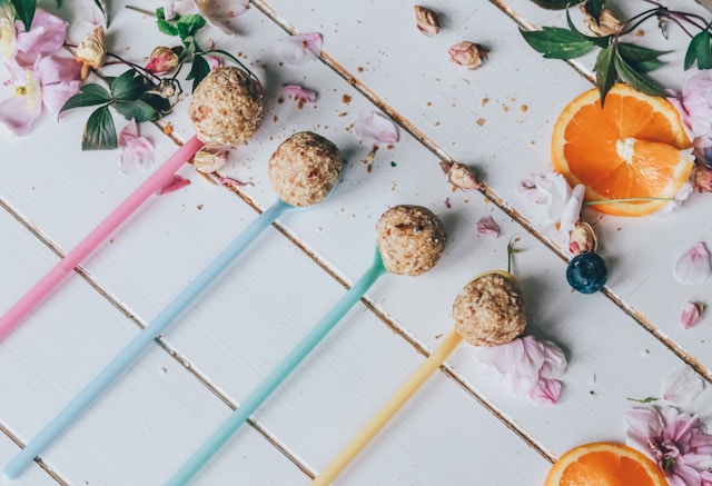 Energy balls on colorful sticks, displayed on a white wooden table with decorative flowers and sliced oranges.