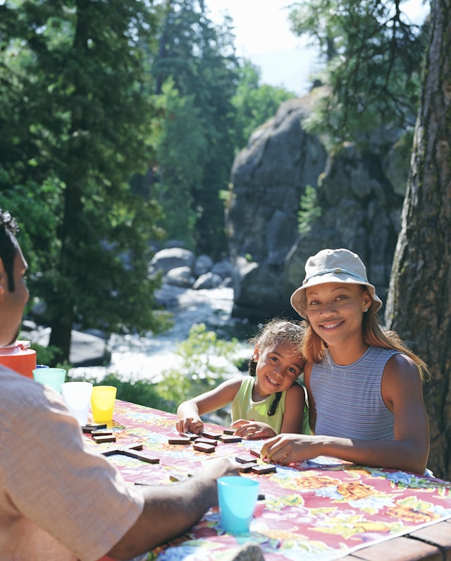 Family playing a board game at a campsite with a scenic river in the background.