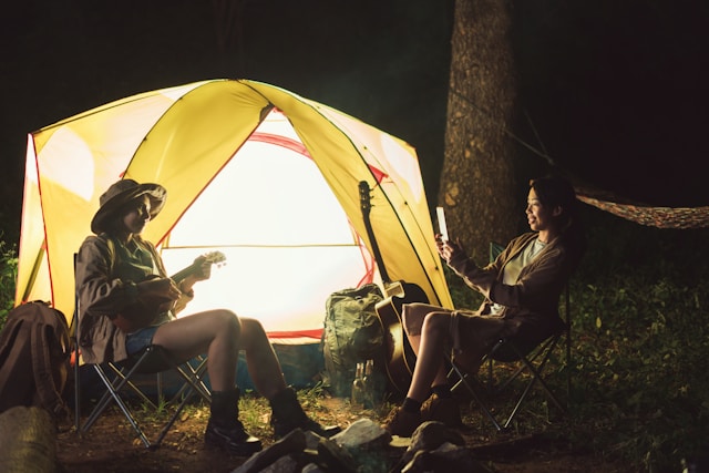 Two campers sitting in front of a tent at night, one playing a musical instrument.