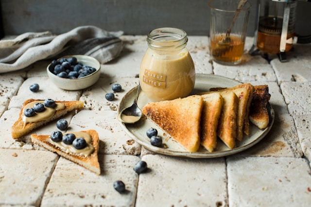 A plate of toast with peanut butter and blueberries, accompanied by a jar of peanut butter and a bowl of fresh blueberries, arranged on a rustic stone surface.