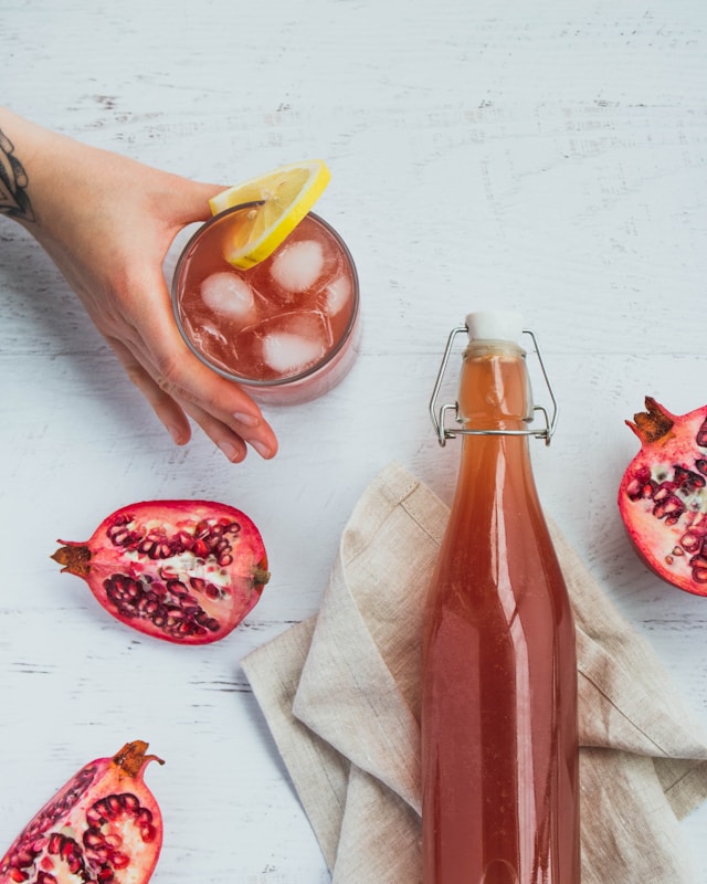 Glass of pomegranate kombucha with lemon and ice, next to a bottle and pomegranate halves