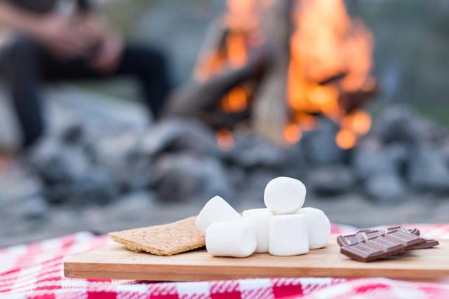 Ingredients for s'mores—marshmallows, chocolate, and graham crackers—on a wooden board with a campfire in the background.