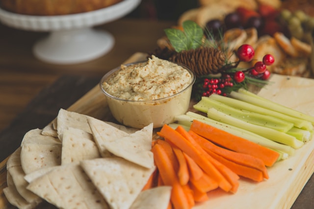 A platter of veggie sticks, including celery and carrots, with pita bread and a bowl of hummus, decorated with holiday-themed greenery and berries.