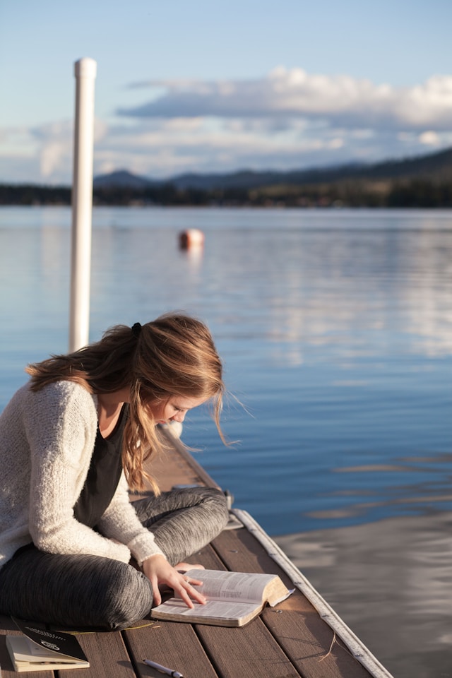 A woman sitting on a dock by the water, reading a book with the scenic view of a lake and mountains in the background.