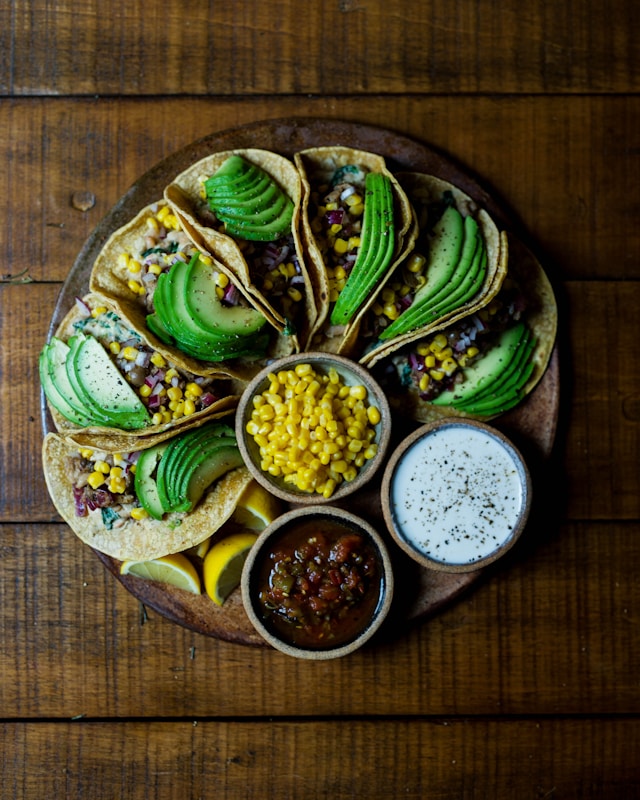 A platter of avocado tacos filled with corn salsa and black beans, accompanied by sides of corn, salsa, and a creamy sauce on a rustic wooden table.