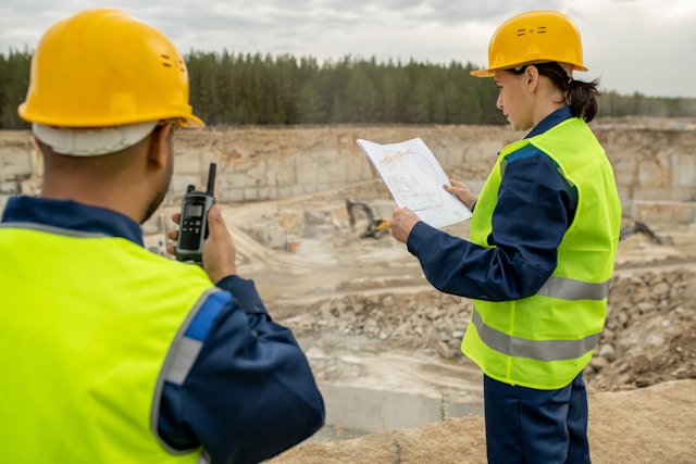 Two construction workers at a quarry, one holding plans and the other speaking into a walkie talkie, both wearing hard hats and safety vests.