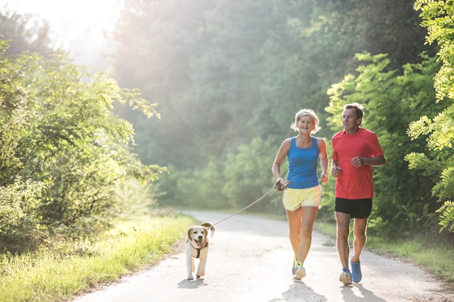 A middle-aged couple, the woman in blue and the man in red, jogging on a forest trail with their small dog on a sunny day.