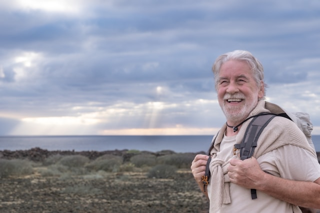 An elderly man with a grey beard, smiling broadly, wearing a backpack, standing against a backdrop of a cloudy sky and calm ocean.