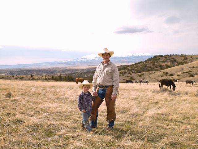 A father and his young son standing in a vast open field with mountains in the background, both dressed in cowboy attire.