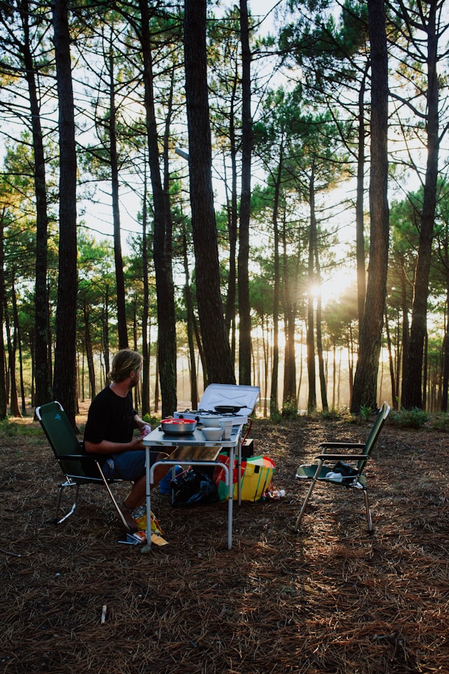 A person sitting at a campsite table surrounded by tall pine trees during sunset.