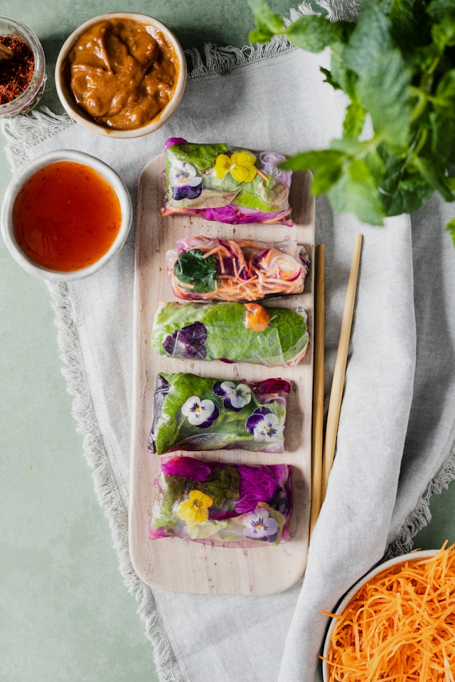 Colorful fresh spring rolls filled with vegetables and edible flowers, served with peanut sauce and sweet chili sauce on a wooden board.