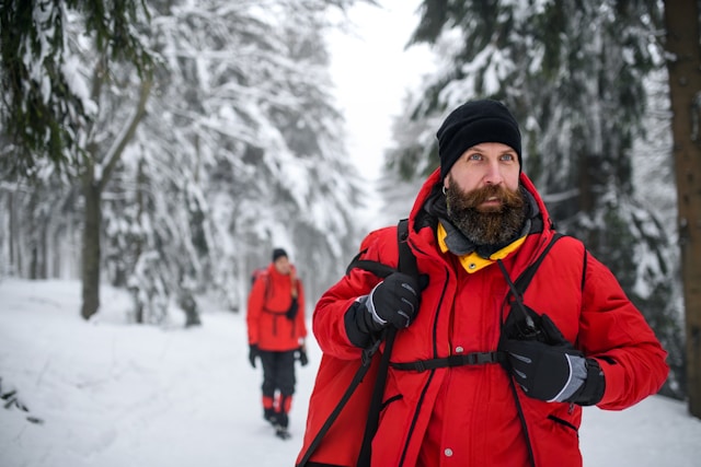 Two hikers in a snowy forest, one in the foreground using a walkie talkie, dressed in a red jacket and black beanie, with another hiker in the background.