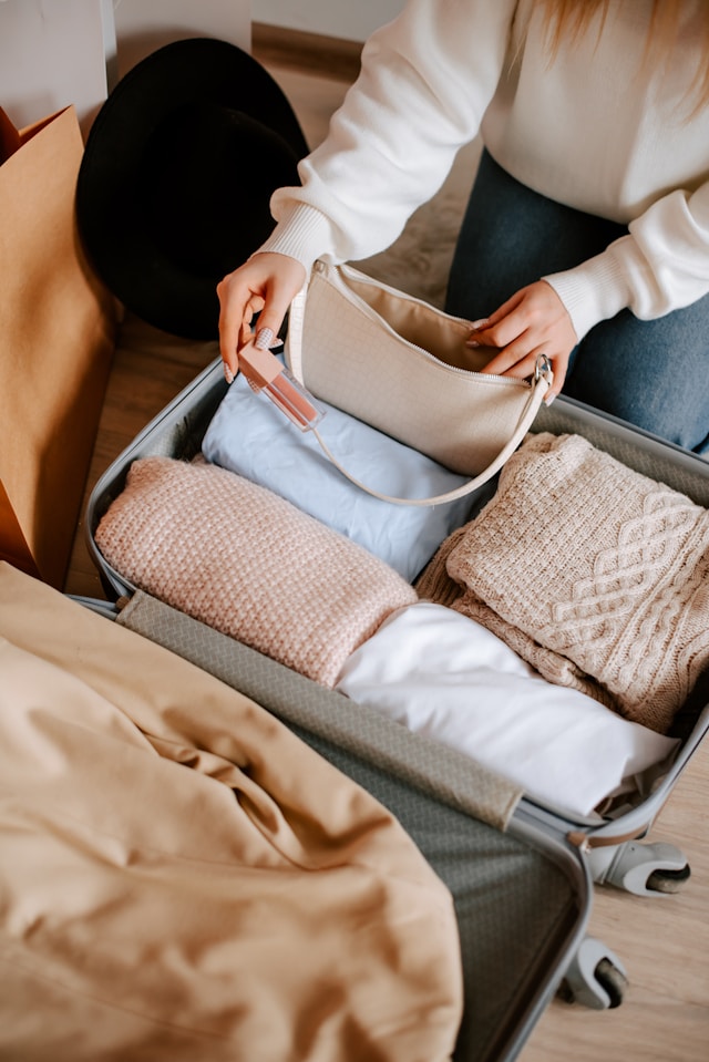 Person packing a suitcase with folded winter clothes and a beige handbag, preparing for a trip.