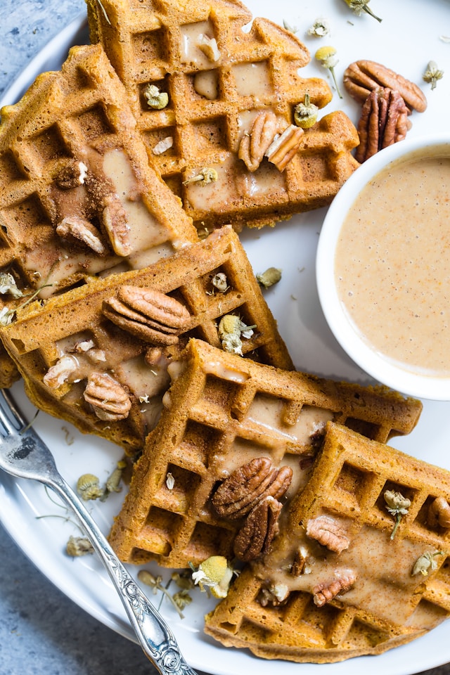 Pumpkin spice waffles topped with pecans and a drizzle of syrup on a white plate with a cup of sauce.