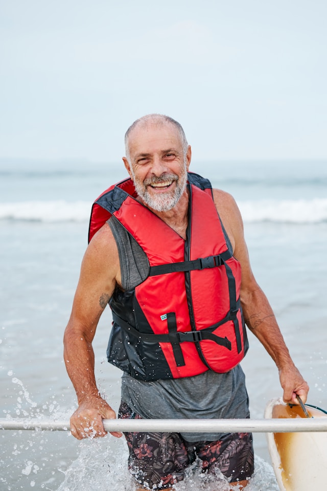 A senior man with a grey beard, wearing a red life jacket, happily kayaking in the sea, water splashing around.