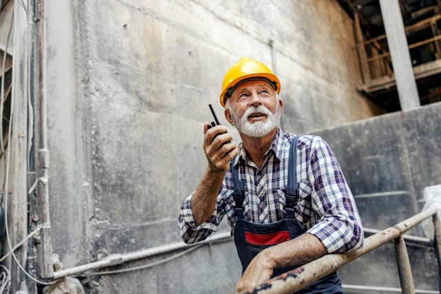 Senior construction worker using a walkie talkie at a construction site, wearing a hard hat and tool belt, looking upward.