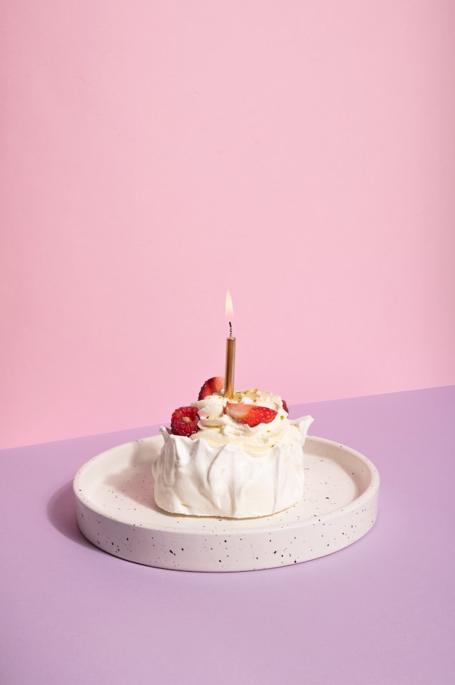 A small birthday cake topped with fresh strawberries and whipped cream, featuring a single lit candle, set against a soft pink background.