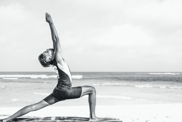 Man performing Warrior Pose on a beach, stretching upwards and looking towards the sky.