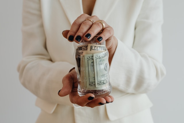 A woman in a white blazer holding a glass jar filled with US dollar bills, showcasing financial savings or donations.