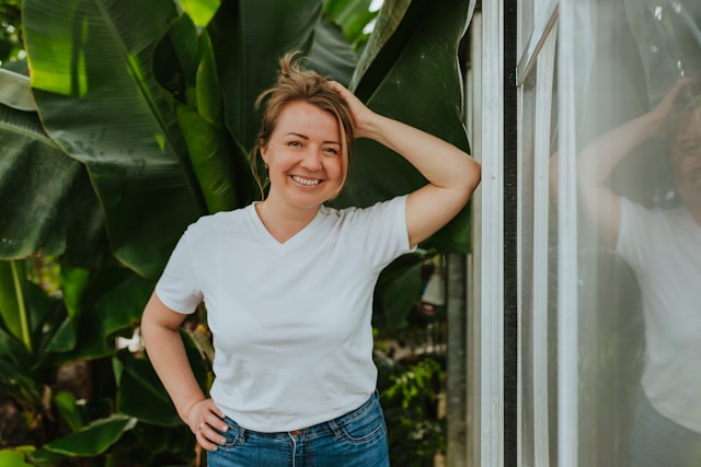 A smiling woman in a white T-shirt and blue jeans standing by a glass door, with large green tropical leaves in the background.