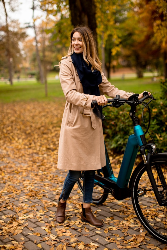 Woman in a trench coat standing with her ebike in an autumn park, surrounded by fallen leaves.