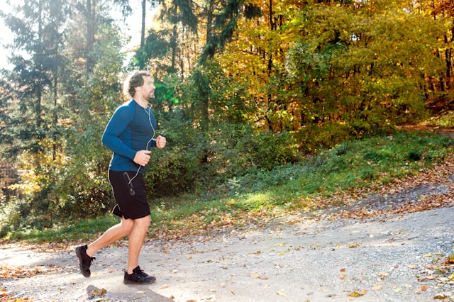 A male runner jogging on a leaf-covered trail in a forest during autumn, representing an active lifestyle amidst nature.